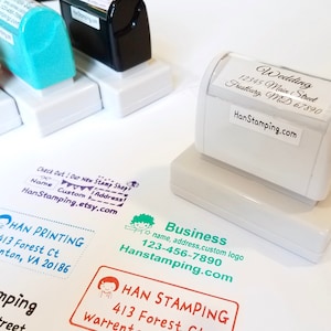 May include: A white self-inking rubber stamp with a label reading Wedding, 12345 Main Street, Frostburg, MD 67890 and HanStamping.com. Nearby are red, teal, and black stamps and various ink impressions with names, addresses, and business logos.