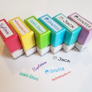 May include: A collection of colorful name stamps in pink, yellow, green, blue, turquoise, and purple. Each stamp has a white label with a name printed in a matching color. The names include "Sylvia", "Isabella Gilmore", "Sawyer", and "Jack".