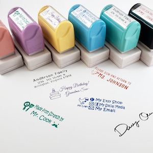 May include: A set of ten colorful self-inking stamps with various designs and text. The stamps are arranged in two rows on a white surface. The text on the stamps includes "Happy Birthday! Grandma Sue", "Made from Scratch by Mr. Cook", "My Etsy Shop", "My Social Media", "My Email", "Anderson Family 1234 5th Street N. Arlington, Virginia 67898", and "Please Sign and Return to MS. JOHNSON".