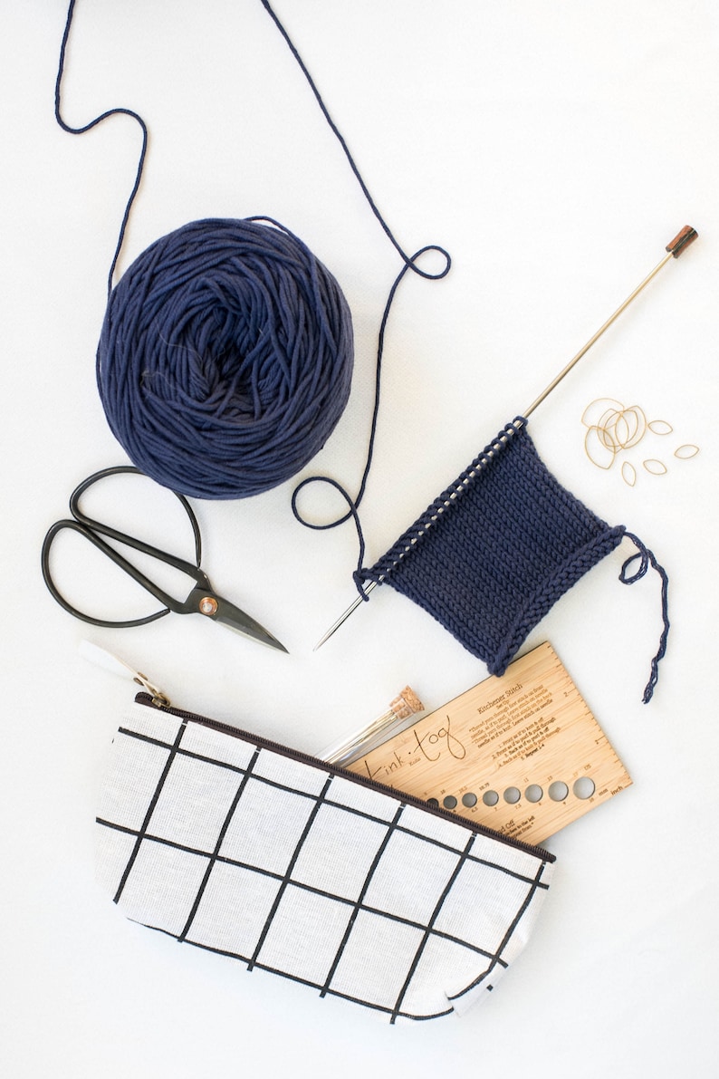 K&ouml;nnte beinhalten: Eine Sammlung von Strickzubeh&ouml;r, darunter ein Kn&auml;uel marineblaues Garn, eine Schere, eine Stricknadel mit einem teilweise gestrickten Quadrat und ein Federm&auml;ppchen mit Gittermuster. Ebenfalls enthalten sind Maschenmarkierer und eine Holzlehre.