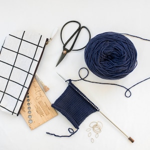 May include: A collection of knitting supplies on a white surface. Includes a navy blue yarn ball, a partially knitted swatch, black scissors, a pencil case with a black grid pattern, and a wooden knitting gauge tool.