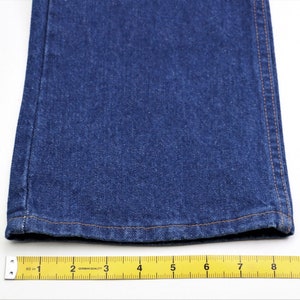May include: Close-up of the bottom hem of a pair of blue denim jeans. The hem is sewn with a light brown thread. A yellow measuring tape is placed below the hem, showing measurements in inches.