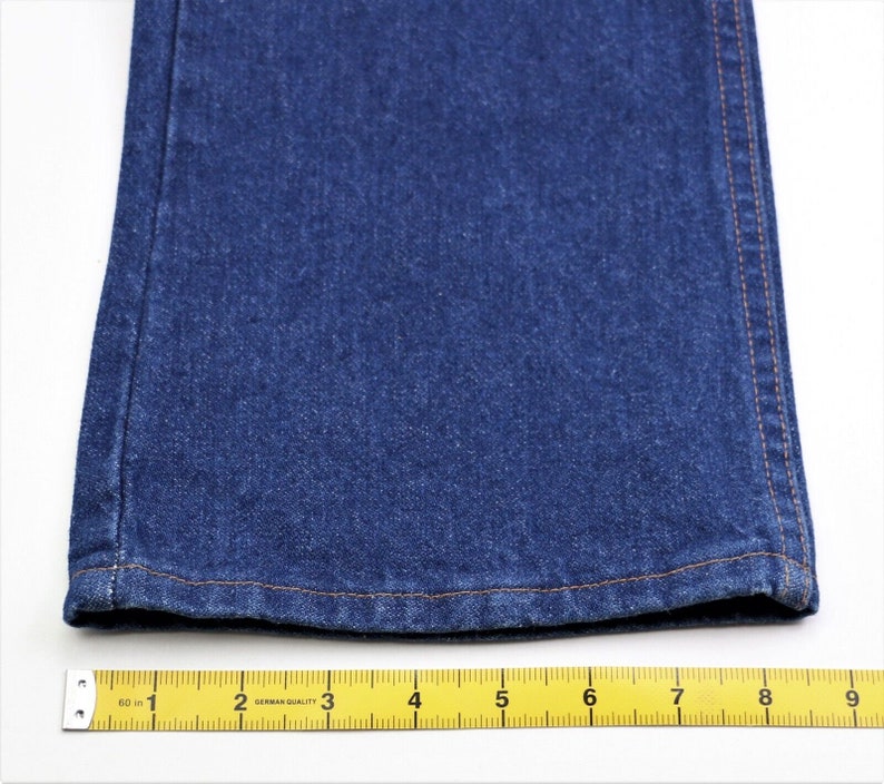 May include: Close-up of the bottom hem of a pair of blue denim jeans. The hem is sewn with a light brown thread. A yellow measuring tape is placed below the hem, showing measurements in inches.