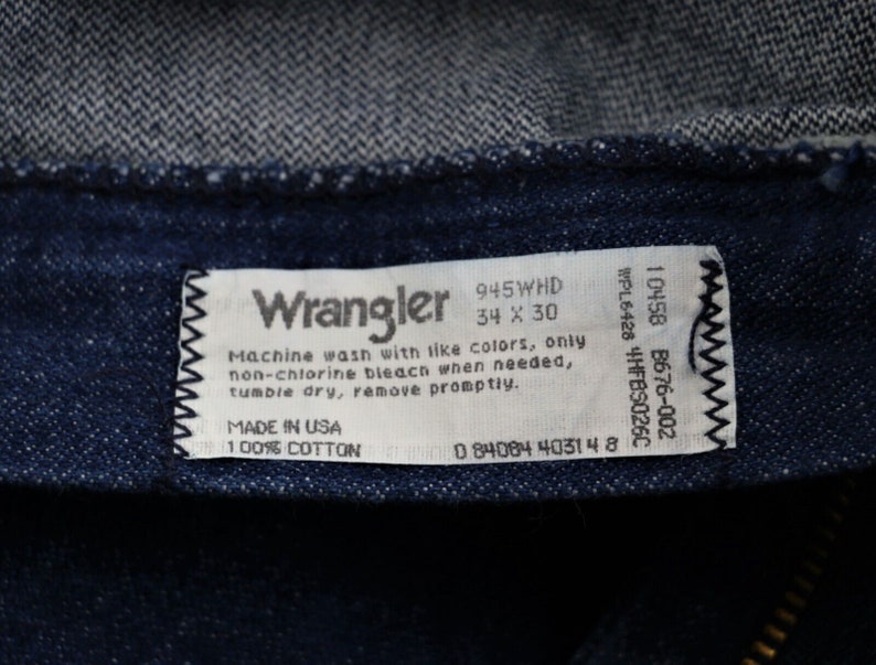 May include: A white Wrangler jeans care label with black text. The label includes the size 34 x 30, the style number 945WHD, and the care instructions: Machine wash with like colors, only non-chlorine bleach when needed, tumble dry, remove promptly. The label also states Made in USA and 100% Cotton.