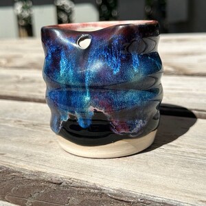 May include: A small, dark blue and black ceramic planter with a white rim and a hole in the side. The planter has a wavy, textured surface and a glossy finish.