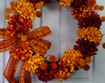 May include: A circular wreath featuring artificial chrysanthemums in shades of deep red, burnt orange, and golden yellow. Small orange pom-pom accents are interspersed throughout the floral arrangement, which is finished with a large plaid ribbon.