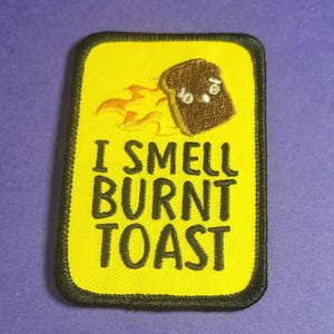 Könnte beinhalten: Gestickter Aufnäher mit gelbem Hintergrund und schwarzem Rand. Der Aufnäher zeigt ein Cartoon-Bild von verbranntem Toast mit Flammen und dem Text "I SMELL BURNT TOAST" in schwarzen Buchstaben.