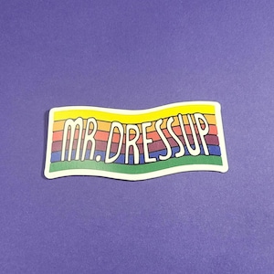 Puede incluir: Una pegatina de colores arcoíris con el texto "MR. DRESSUP" en blanco.