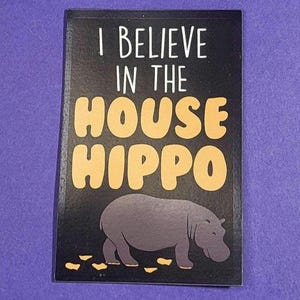 Può includere: Un magnete rettangolare nero con la scritta "I BELIEVE IN THE HOUSE HIPPO" in bianco e giallo. Un'illustrazione di ippopotamo grigio in basso. Il magnete è su una superficie viola.