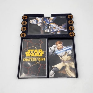 Peut inclure: Un porte-cartes noir sur le thème de Star Wars Shatterpoint. Le porte-cartes comporte trois emplacements pour cartes. L'emplacement supérieur présente une image d'un personnage de Star Wars. L'emplacement du milieu présente le logo de Star Wars Shatterpoint. L'emplacement inférieur présente une image d'un personnage de Star Wars.