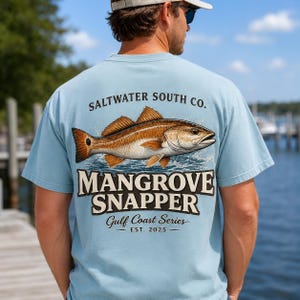 May include: A light blue t-shirt featuring a graphic on the back of a brown and tan fish swimming in water. The text reads SALTWATER SOUTH CO., MANGROVE SNAPPER, Gulf Coast Series, and EST. 2025 in black and dark brown lettering.