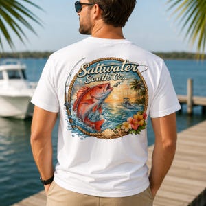 May include: A white t-shirt featuring a circular graphic on the back. The design shows a red fish jumping out of the water, a fishing boat, a palm tree, and hibiscus flowers, framed by a rope border with the text Saltwater South Co.