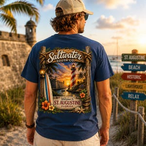 Può includere: Una t-shirt blu navy con una stampa grafica sul retro. Il design mostra un faro, una tavola da surf e fiori tropicali incorniciati da pali di legno e corda. Il testo recita Saltwater South Co. e ST. AUGUSTINE FLORIDA.