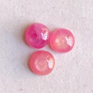 May include: Three small, round, cabochon-cut gemstones in varying shades of pink. Each stone features a smooth, polished, domed surface that reflects light, displaying a translucent quality with subtle internal variations in color and texture.