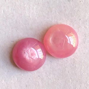 以下が含まれることがあります： 滑らかで磨かれた表面を持つ、2つの小さな丸いカボションカットの宝石。片方は鮮やかなマゼンタピンクで、もう片方はより明るく半透明なコーラルピンクです。どちらの石も光沢のある輝きを放ち、丸みを帯びたドーム状の形状をしています。