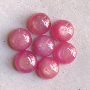 Può includere: Una collezione di sette pietre cabochon rosa, circolari e lisce, disposte a grappolo. Ogni pietra presenta una superficie lucida e brillante con una parte superiore a cupola e una tonalità rosa vibrante e uniforme che riflette la luce.