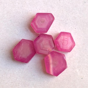 May include: Five small, hexagonal-shaped pink gemstones arranged in a cluster. Each stone features a distinct, layered geometric pattern with concentric hexagonal lines visible within the translucent, vibrant pink material of the polished surfaces.