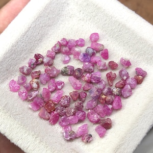 Può includere: Una collezione di piccole gemme di rubino grezze in varie tonalità di rosa e rosso. Le gemme sono sparse su una superficie bianca e strutturata, mostrando le loro forme naturali e irregolari e le sottili variazioni di colore.