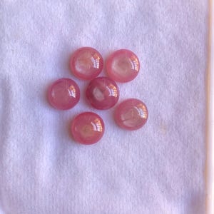 Può includere: Sei gemme rosa rotonde e lucide sono disposte in un vassoio bianco. Le gemme hanno una qualità traslucida, con un centro più chiaro, quasi giallo. Il vassoio è foderato con un tessuto bianco strutturato.