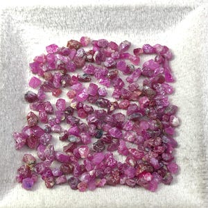 Puede incluir: Una colección de pequeñas piedras preciosas de rubí en bruto. Las gemas varían en tamaño y forma, mostrando tonos de rosa y rojo. Se encuentran dispersas sobre una superficie blanca y texturizada, creando una exhibición visualmente atractiva de piedras preciosas naturales.