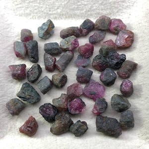 Peut inclure: Une collection de pierres précieuses de rubis brutes de différentes tailles et formes. Les pierres présentent une gamme de couleurs, notamment des rouges profonds, des roses et des violets, avec quelques reflets gris et bleus. Les pierres sont dispersées sur une surface blanche texturée.