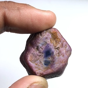 May include: A raw, uncut gemstone with a hexagonal shape, featuring a mix of deep magenta and purple hues. The surface has a textured, natural appearance with visible brown inclusions and a dark, concentrated blue-purple center point.