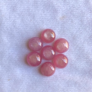 Peut inclure: Sept pierres précieuses roses et rondes, avec une finition polie et brillante, sont disposées sur une surface blanche et texturée. Les gemmes ont une qualité translucide, reflétant la lumière et créant des reflets. Les pierres sont probablement destinées à la joaillerie.