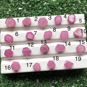 May include: A collection of nineteen pink gemstones, each with a unique hexagonal or irregular shape, arranged on a white display stand. The gemstones vary in size and shade, with some appearing more translucent than others. The background is a bed of green, leafy material.