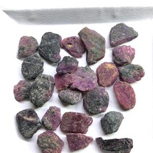 Op de afbeelding: Een verzameling ruw geslepen robijn edelstenen in verschillende vormen en maten. De stenen vertonen een scala aan kleuren, van diep rood en paarsrood tot grijs en zwart, met een ruw, getextureerd oppervlak. De edelstenen zijn verspreid op een wit oppervlak.