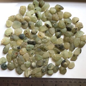 May include: A collection of small, tumbled pale green stones with dark inclusions, scattered above a metal ruler that shows measurements in centimetres and inches, with the stones ranging in size from approximately 0.5 to 2 cm in diameter.