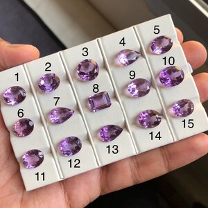 May include: A selection of fifteen amethyst gemstones in various shapes, including oval, pear, and emerald cuts. The gemstones are displayed on a white tray, showcasing their vibrant purple hues. The gemstones are numbered 1 through 15.