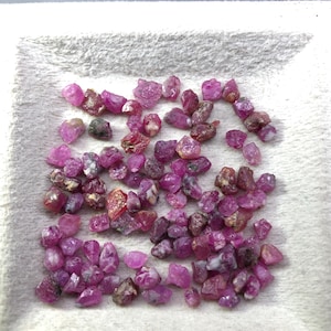 Peut inclure: Une collection de petits rubis bruts de couleur rose et rouge. Les pierres précieuses sont dispersées sur une surface blanche et texturée, mettant en valeur leurs formes naturelles irrégulières et leur translucidité variable.