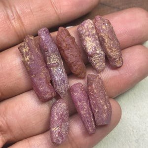 Op de afbeelding: Een verzameling ruwe, ongeslepen robijnkristallen. De edelstenen variëren in grootte en vorm en vertonen een dieprode en paarsrode tint, met enkele lichtere gebieden. De kristallen hebben een natuurlijke, ongepolijste uitstraling.