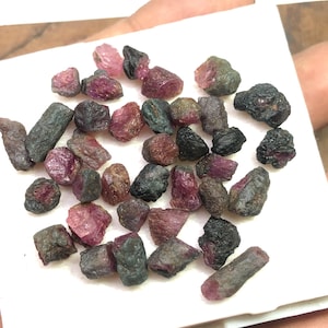 Könnte beinhalten: Eine Sammlung roher, ungeschliffener Turmalin-Edelsteine in verschiedenen Rosa-, Grün- und Schwarztönen. Die Edelsteine werden auf einer weißen Oberfläche präsentiert, die ihre natürlichen, rauen Texturen und Farben hervorhebt. Ideal für Schmuckherstellung oder Sammlungen.