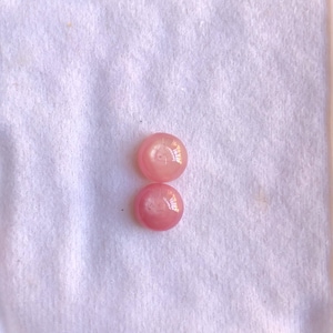 Può includere: Due perle rosa rotonde e lucenti sono esposte su una superficie bianca e strutturata. Le perle sono racchiuse in un contenitore bianco, suggerendo che facciano parte di una collezione di gioielli o di un accessorio artigianale.