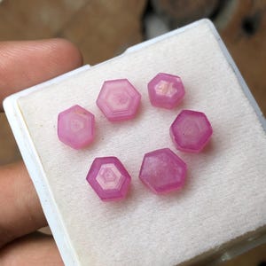 May include: A collection of six hexagonal, pink gemstones displayed on a white surface. Each gem has a layered, concentric design within its structure. The gems are set in a white, rectangular container.