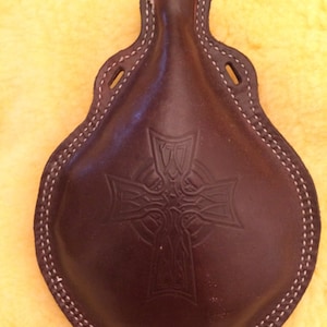 May include: A brown leather flask with a wooden top and a Celtic cross design. The flask has white stitching around the edges.