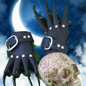 May include: A pair of black fingerless gloves featuring long, curved, claw-like extensions on each finger, secured with silver-toned metal rivets and adjustable wrist straps with large silver buckles, positioned next to a decorative human skull.