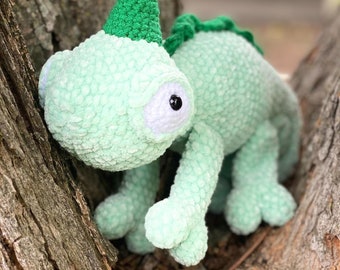 May include: A green crocheted chameleon plush toy with large black eyes and a green crocheted hat. The chameleon is perched on a tree branch.