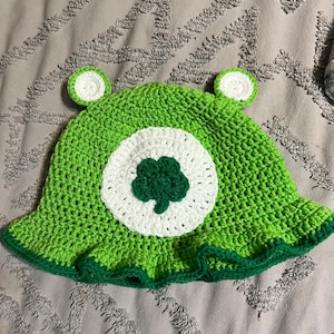 May include: Green crocheted hat with a white clover design and green ears.