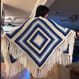 Puede incluir: Un chal cuadrado de ganchillo con un patrón de diamantes en tonos azul marino, azul claro y crema. La prenda está confeccionada con un diseño de punto granny square clásico y terminada con largos flecos de color crema en todos los bordes.