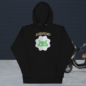 Puede incluir: Una sudadera negra con capucha y un gráfico en el pecho. El gráfico muestra la palabra ANDROID en letras metálicas sobre un emblema circular blanco de borde festoneado que contiene un icono de cabeza de robot Android verde claro.