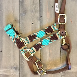 May include: A brown and turquoise horse halter with gold-colored hardware. The halter features a woven design with turquoise diamond shapes and brown and tan accents. The hardware includes buckles and a snap hook. The halter is designed for equine use.