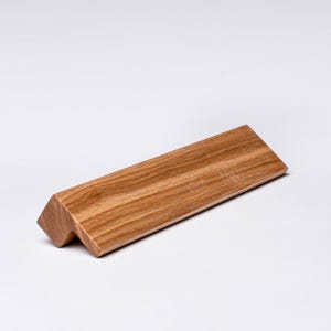 Pode incluir: Um pequeno bloco de madeira angular com um acabamento natural de grão de madeira. O bloco é retangular com uma borda chanfrada e parece ser um acessório de mesa ou um item decorativo. A madeira tem uma cor castanha clara e quente.