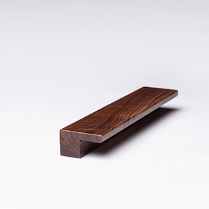 以下が含まれることがあります： 長方形で、端がわずかに角度のついた、濃い茶色の木製キャビネットプルです。