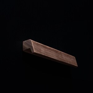 Puede incluir: Un tirador de armario rectangular de madera con borde biselado. El tirador es de color marrón cálido y está sobre un fondo negro. Este herraje es adecuado para cajones y armarios.