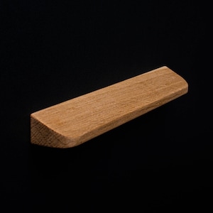 May include: A small, rectangular wooden object with a natural wood grain finish. One end is cut at an angle, and the other end is rounded. The object is set against a solid black background.