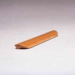 May include: A light brown, wooden door stop with a rounded end and a curved, angled edge. The door stop is made of wood and has visible grain lines. The door stop is approximately 8 inches long.