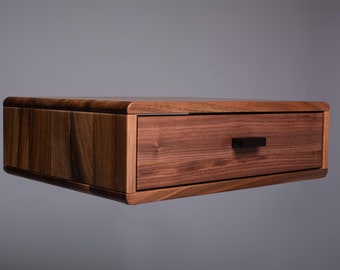 Pode incluir: Uma mesa de cabeceira flutuante de madeira de noz com uma única gaveta e uma puxador de metal preto. A mesa de cabeceira tem uma borda curva e um acabamento de madeira natural.