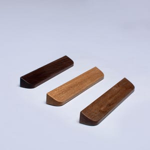 Peut inclure: Trois poignées de tiroir en bois dans différentes teintes de bois. Les poignées sont toutes rectangulaires avec des bords arrondis. Les poignées sont disposées en rangée de gauche à droite, avec la poignée en bois la plus foncée à gauche, la poignée en bois la plus claire au milieu et la poignée en bois moyenne à droite.